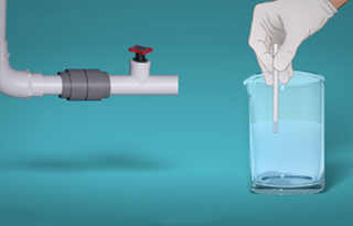 Gloved hand inserting a tool into a beaker of water next to a pipe valve assembly on a teal background